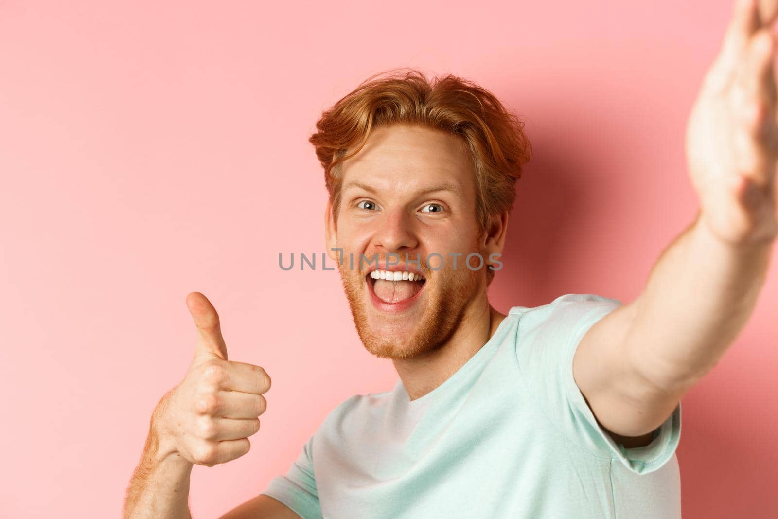 Excited redhead man tourist taking selfie and showing thumbs-up, holding camera with stretched out hand, standing over pink background by Benzoix