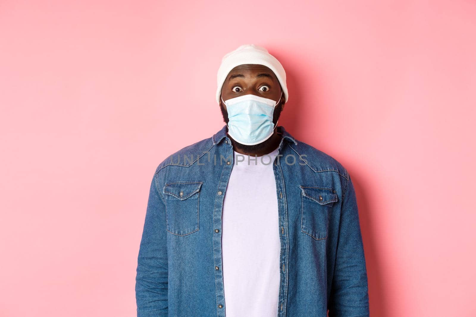 Covid-19, lifestyle and quarantine concept. Image of impressed african-american man in face mask staring at camera, gasping surprised, standing over pink background by Benzoix