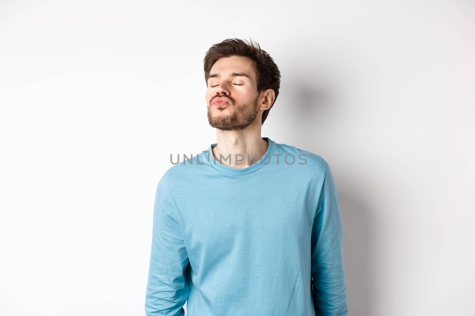 Image of cute and silly man waiting for kiss, pucker lips with closed eyes, standing over white background.