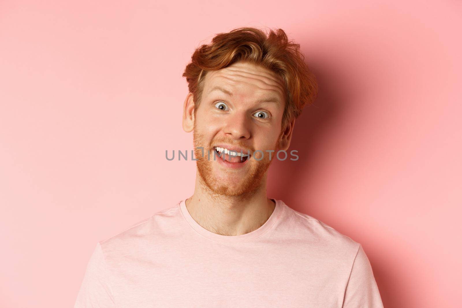 Headshot of funny redhead guy showing tongue, making silly faces at camera, standing joyful against pink background. Copy space