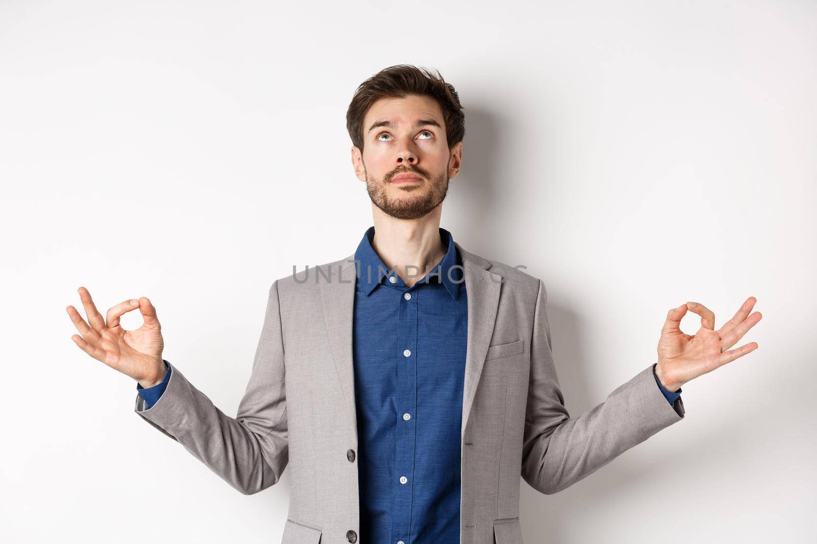 Calm ceo manager in suit relaxing with meditation, looking up and holding hands in zen mudra sign, standing on white background.