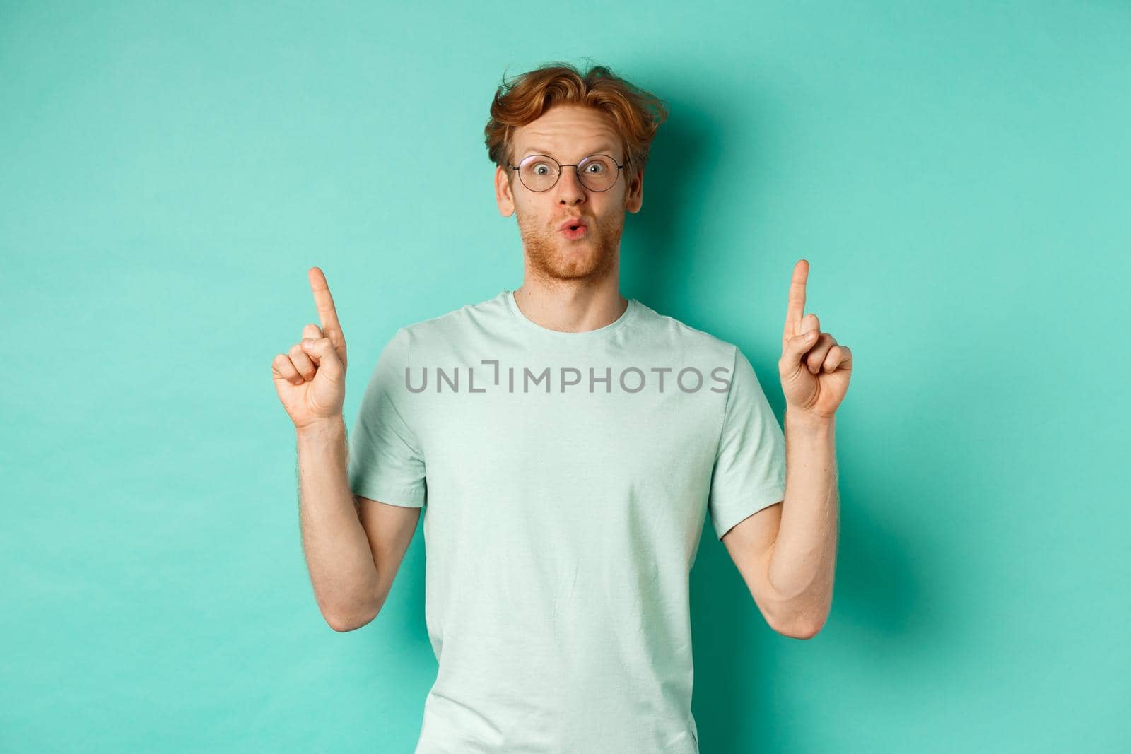 Impressed redhead man in glasses and t-shirt, checking out promo offer, pointing fingers up at copy space, staring at camera amazed, standing over turquoise background.