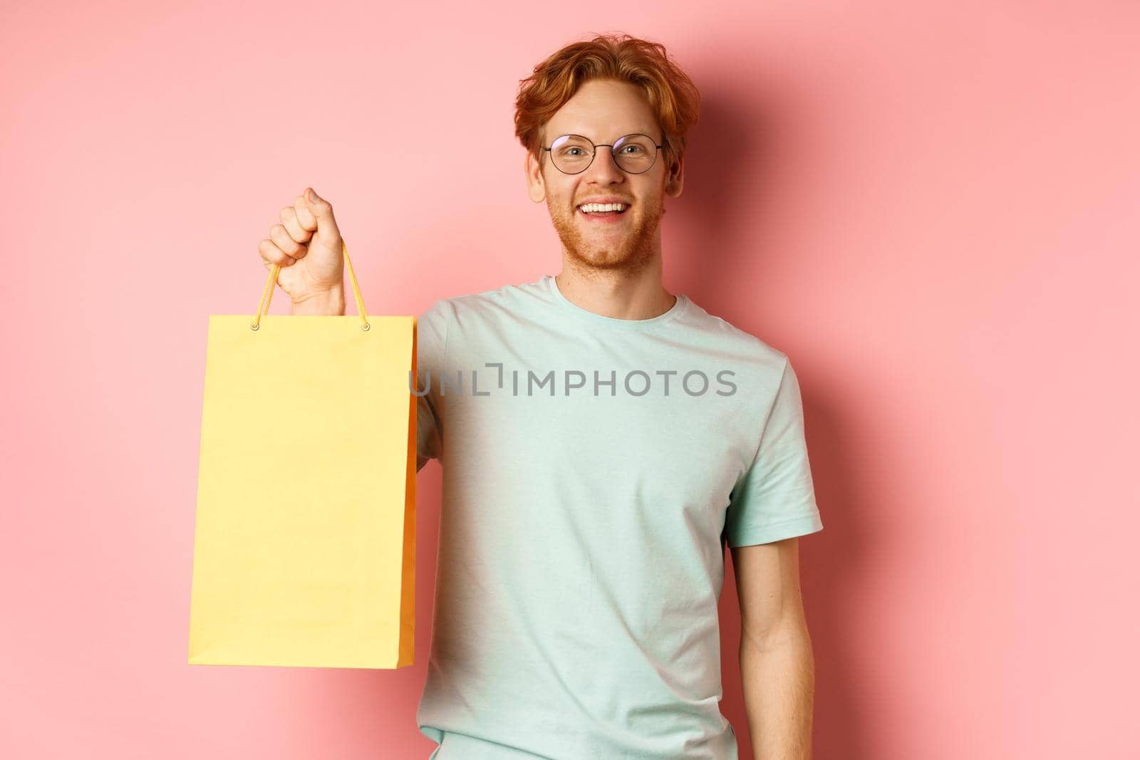 Handsome young man buying presents, holding shopping bag and smiling at camera, standing over pink background by Benzoix