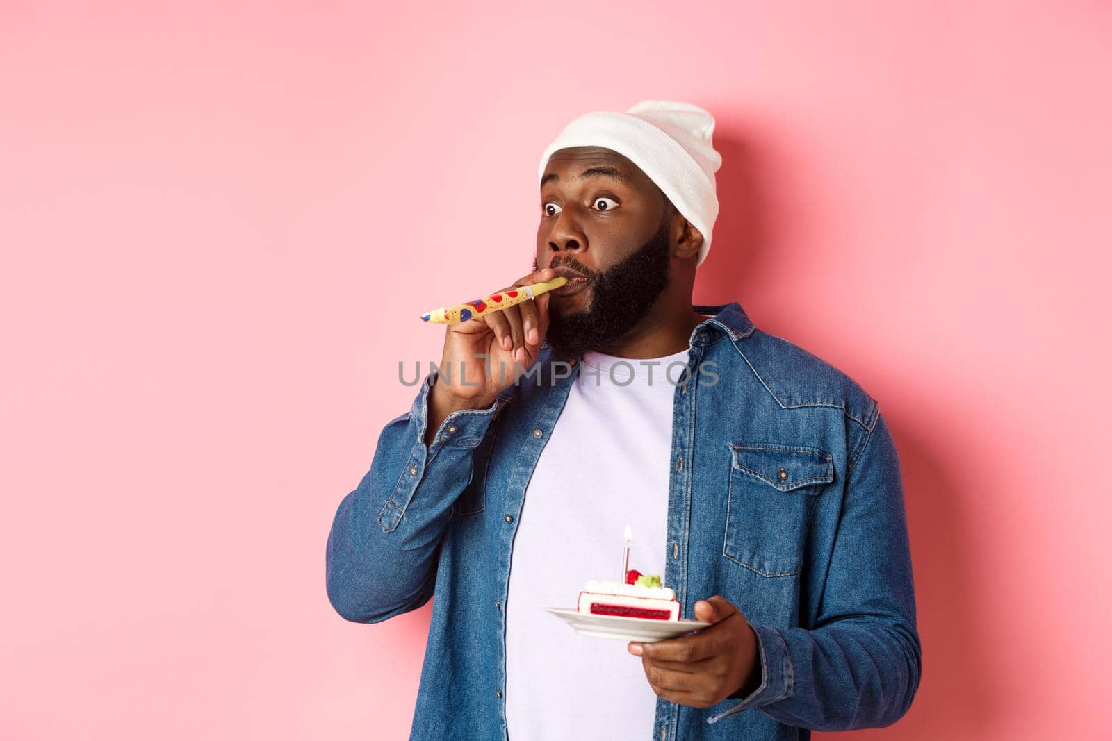 Happy Black hipster celebrating birthday, blowing party whistle, holding bday cake with candle, standing over pink background by Benzoix