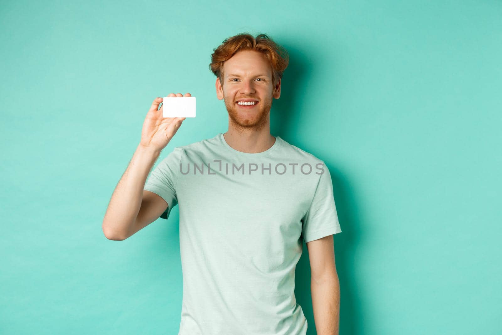 Shopping concept. Handsome redhead man in t-shirt showing plastic credit card and smiling, standing over turquoise background by Benzoix