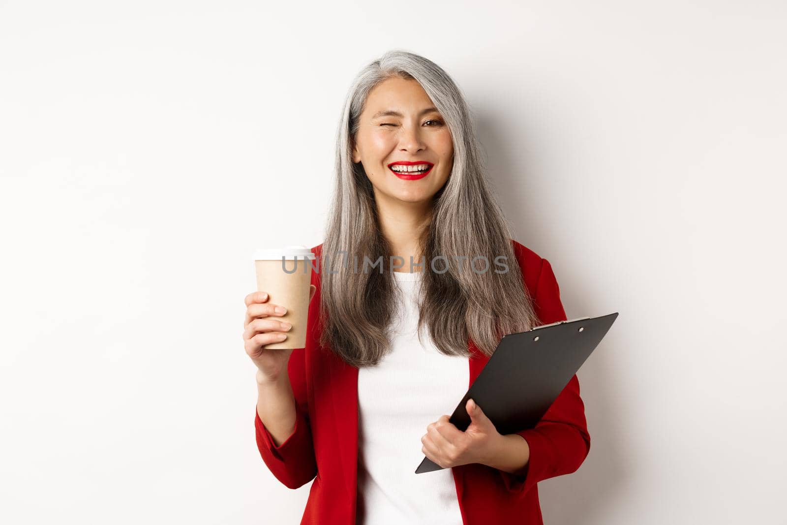 Successful asian businesswoman drinking coffee and winking cheerful at camera, standing with clipboard, wearing red blazer, white background.