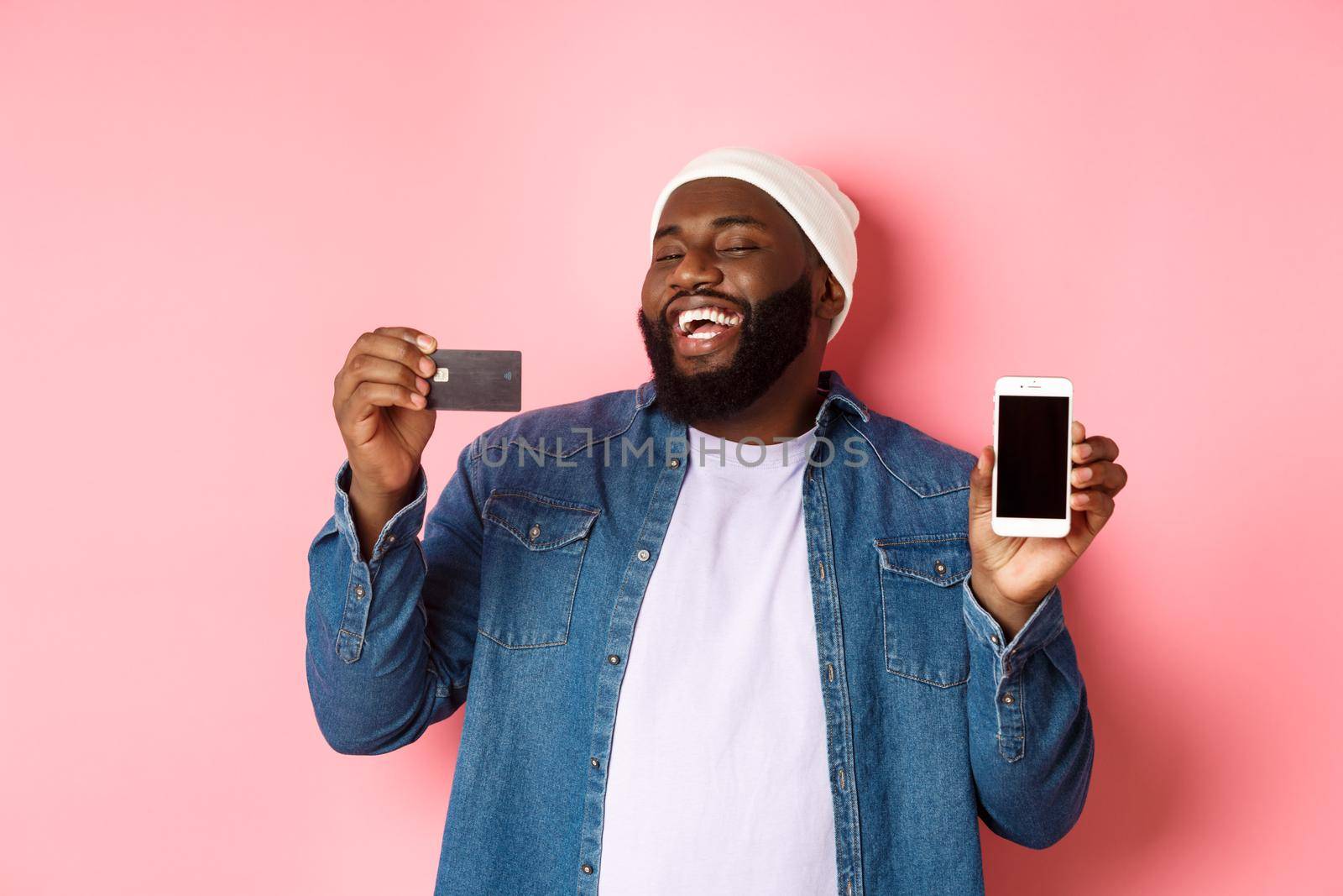 Online shopping. Happy african-american man in beanie laughing, showing credit card and mobile phone screen, standing over pink background by Benzoix