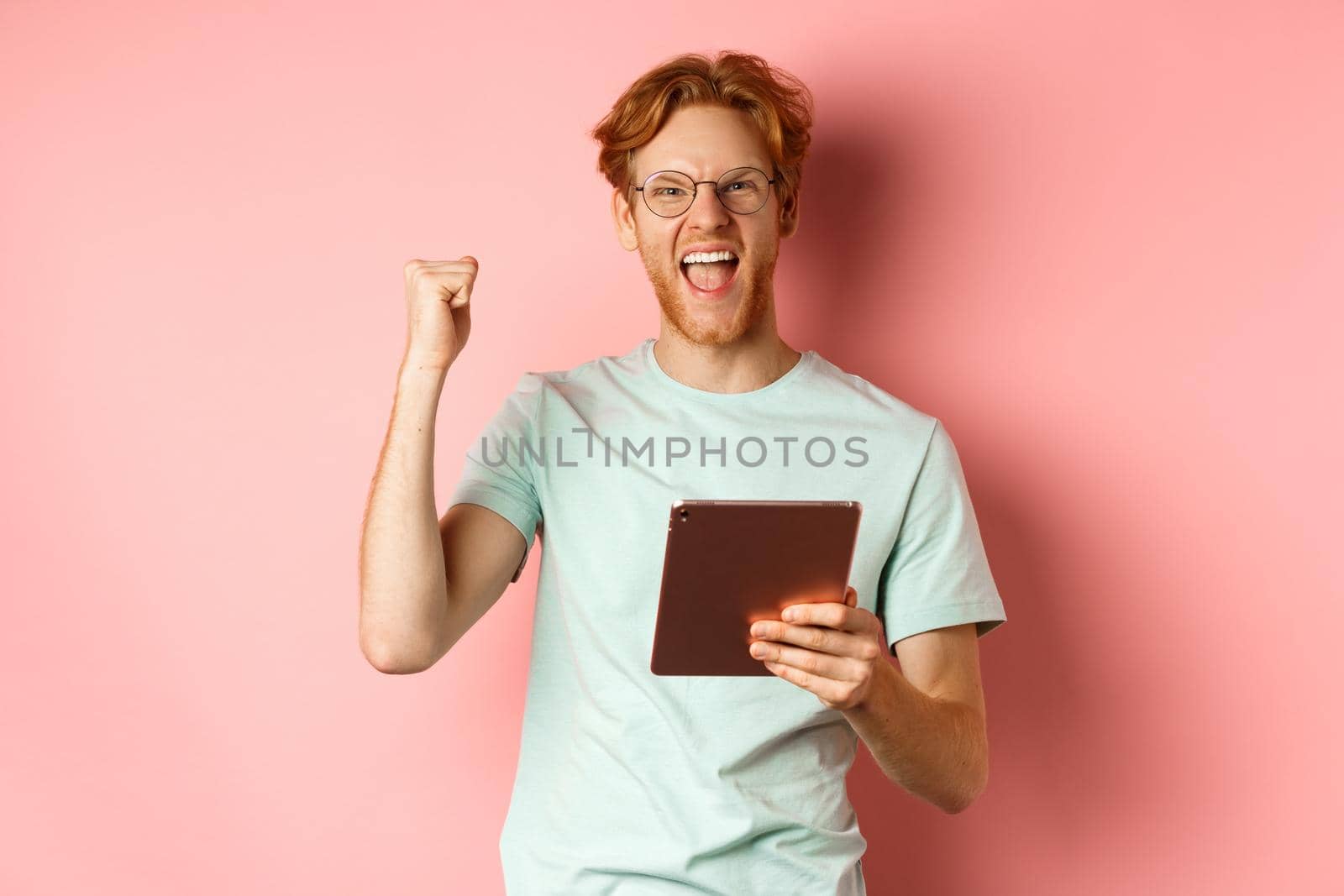 Image of happy redhead man triumphing, winning online with digital tablet and rejoicing, standing over pink background by Benzoix