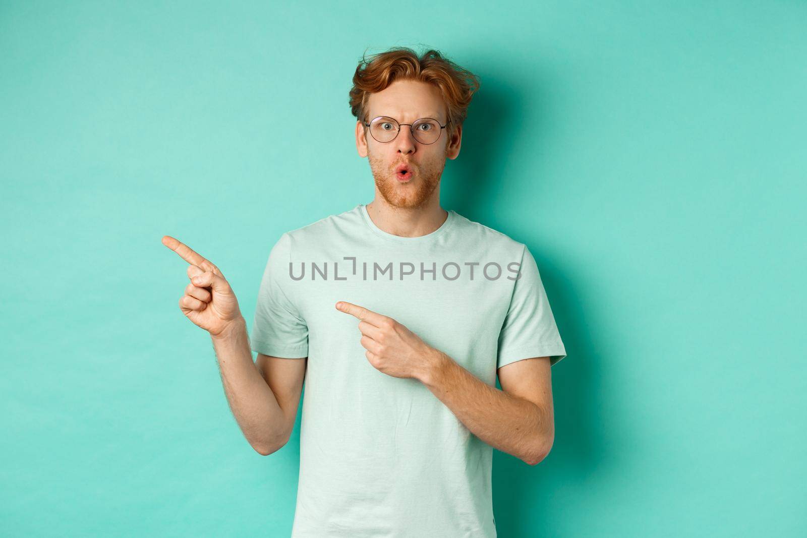 Surprised redhead guy in glasses and t-shirt pointing fingers left, saying wow and showing promo offer, checking out special deal, standing over turquoise background.