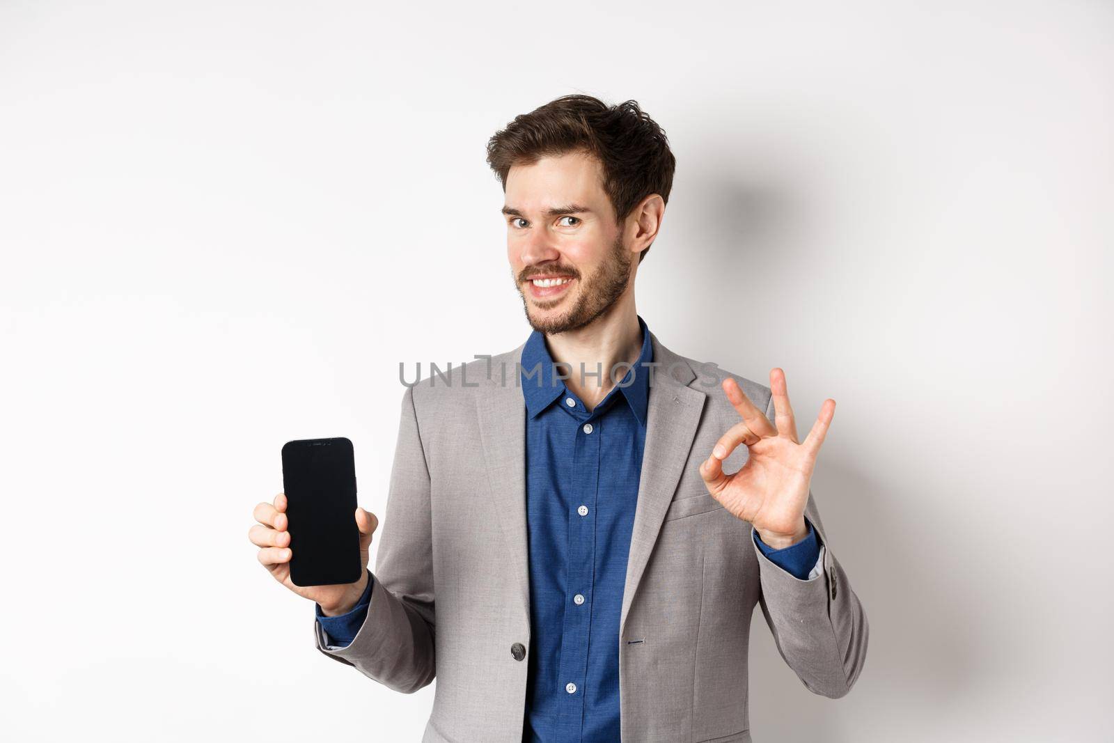 E-commerce and online shopping concept. Smiling businessman show okay sign and empty smartphone screen, demonstrate his account, standing on white background.