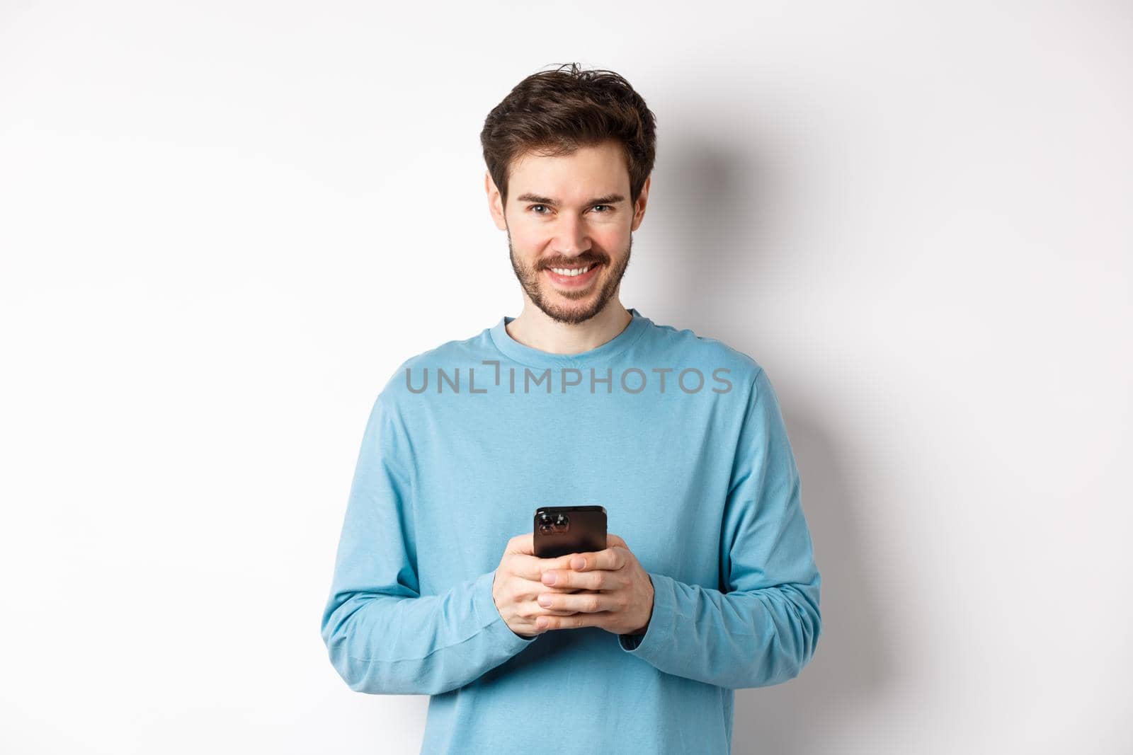 Smiling modern man using smartphone and looking pleased at camera. Guy in sweatshirt with mobile phone on white background by Benzoix