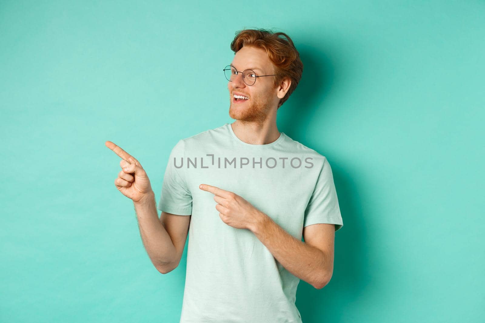 Handsome young man with red hair and beard, wearing glasses and t-shirt, pointing and looking left with amused face, checking out advertisement on copy space, mint background.
