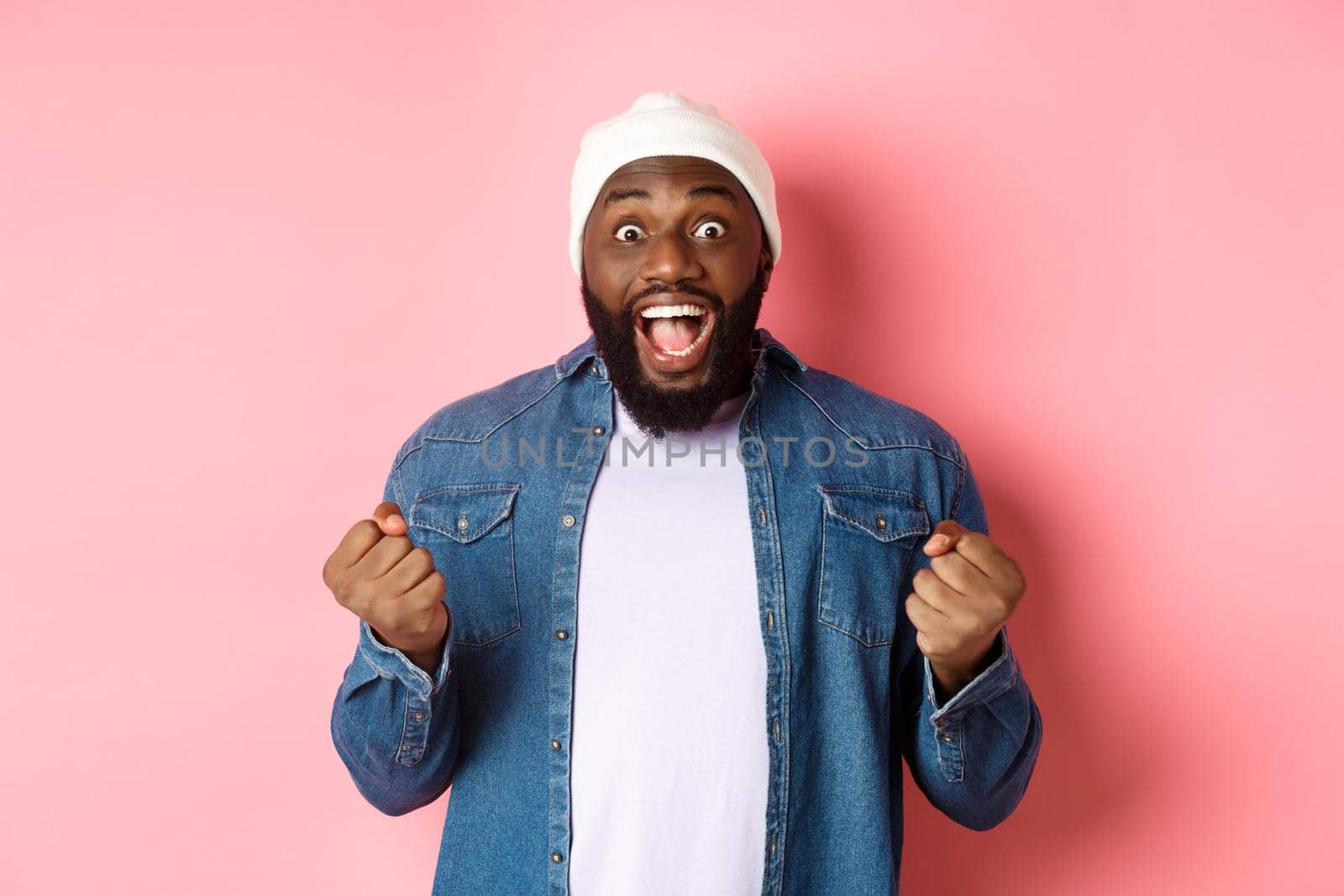 Surprised and happy man staring with disbelief while winning, clench fists and smiling amazed, celebrating victory, achieve goal, standing over pink background by Benzoix