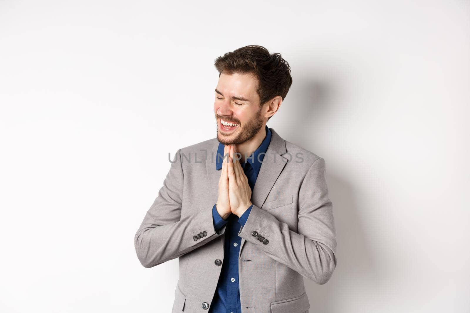 Happy and thankful businessman rejoicing, holding hands in begging or namaste sign, thanking for help, smiling relieved, standing in suit on white background.