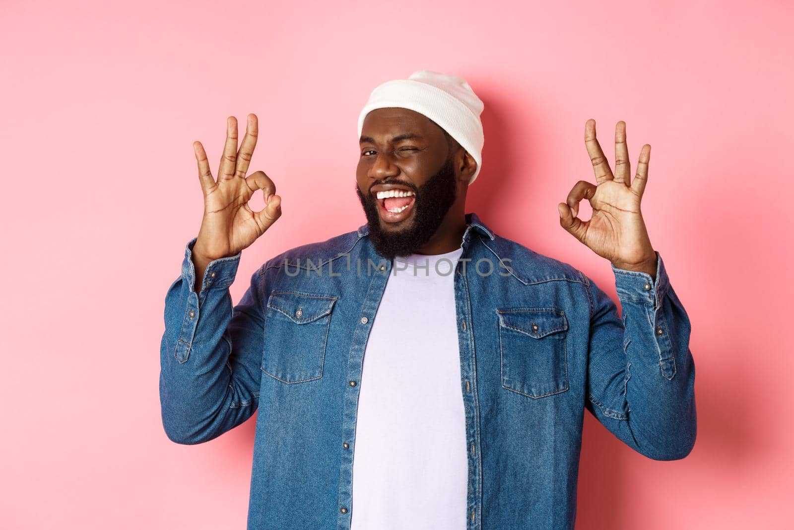 Satisfied african-american man in hipster beanie, showing okay signs in approval, praising good offer and saying yes, smiling pleased, standing over pink background.