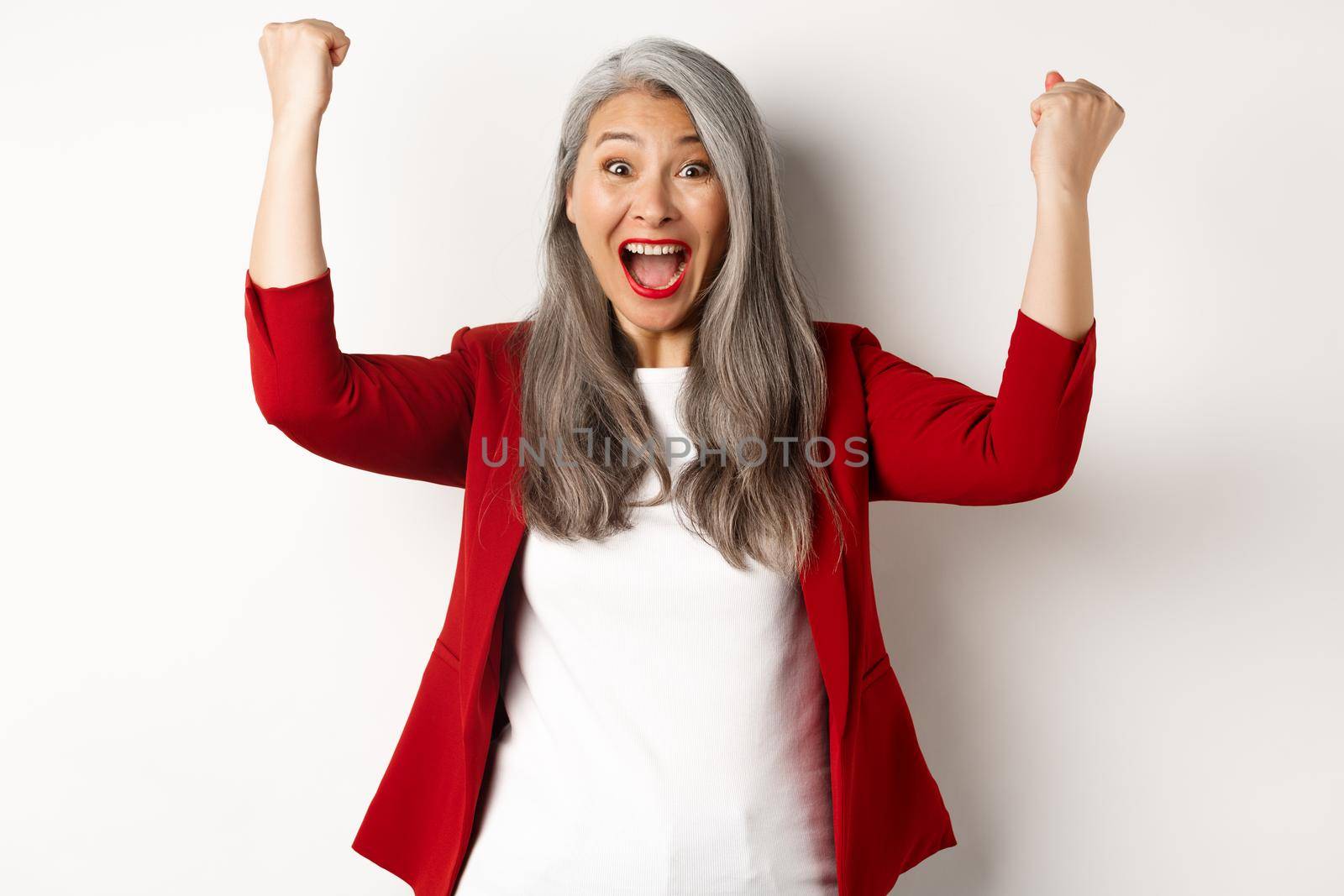 Lucky senior woman achieve success, winning prize and celebrating, saying yes with fist pumps, standing happy against white background.