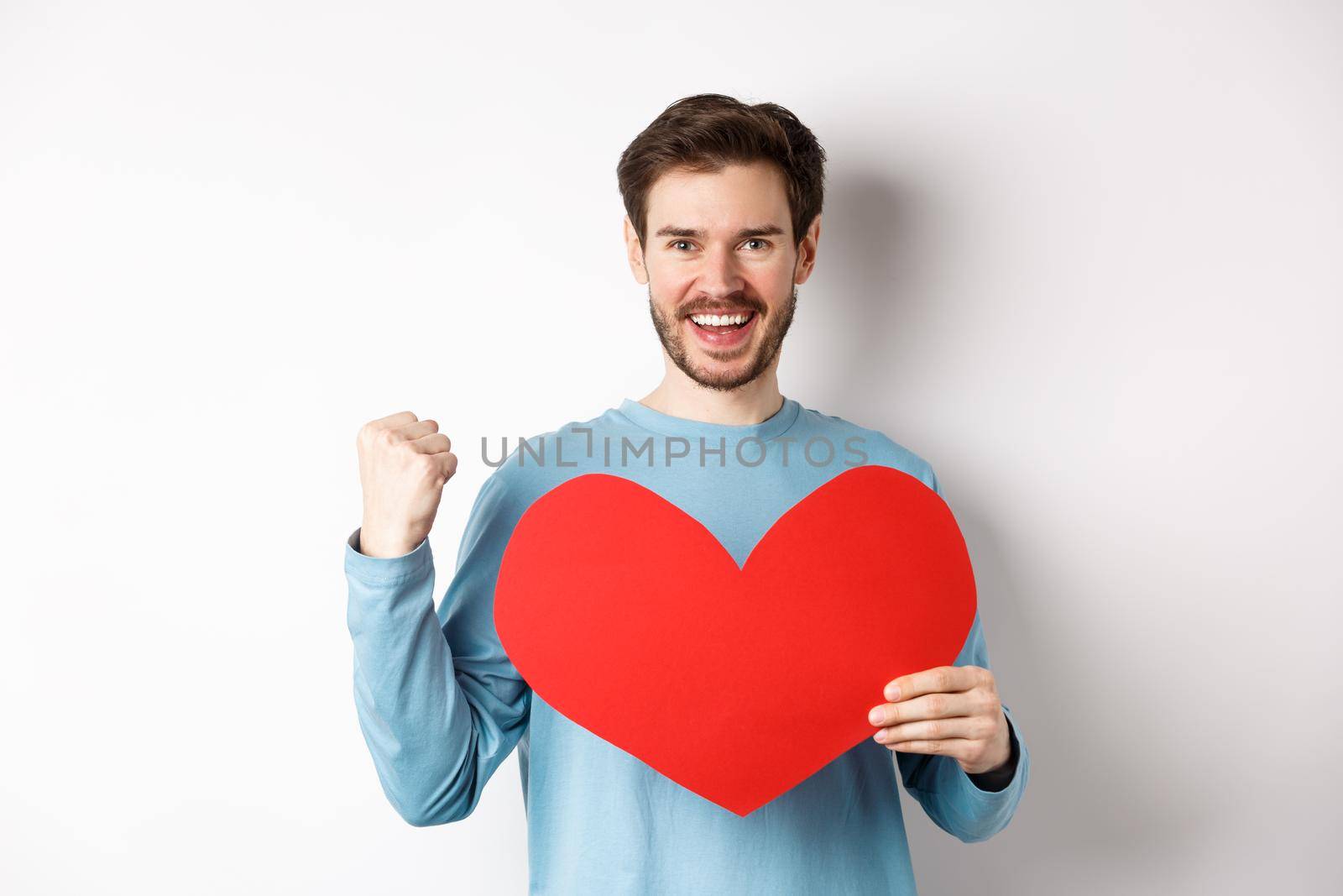 Cheerful man determined to find love on Valentines day, making fist pump success gesture and holding big red romantic heart, standing over white background.
