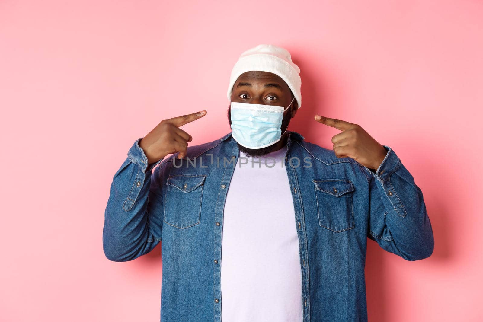 Coronavirus, lifestyle and social distancing concept. Happy Black man in beanie pointing at his face mask, smiling at camera, standing over pink background by Benzoix