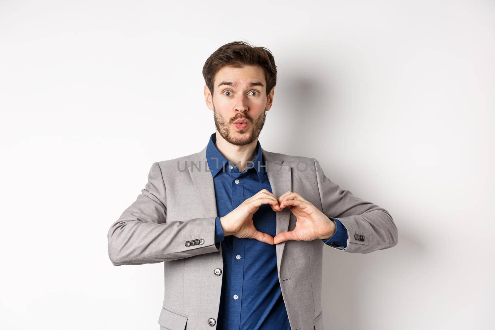 Handsome man looking silly, showing heart love you sign and pucker lips, waiting for kiss from lover, standing in suit on white background by Benzoix