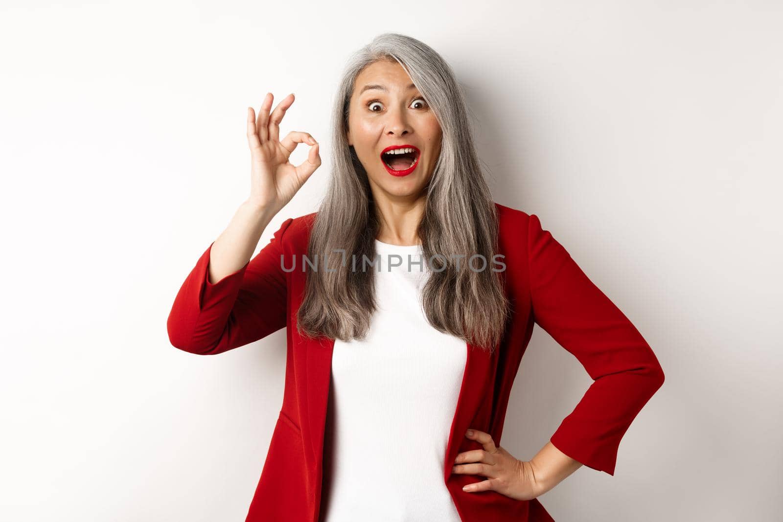 Happy asian mature woman in red blazer showing okay sign, smiling amazed, praising something awesome, standing over white background.