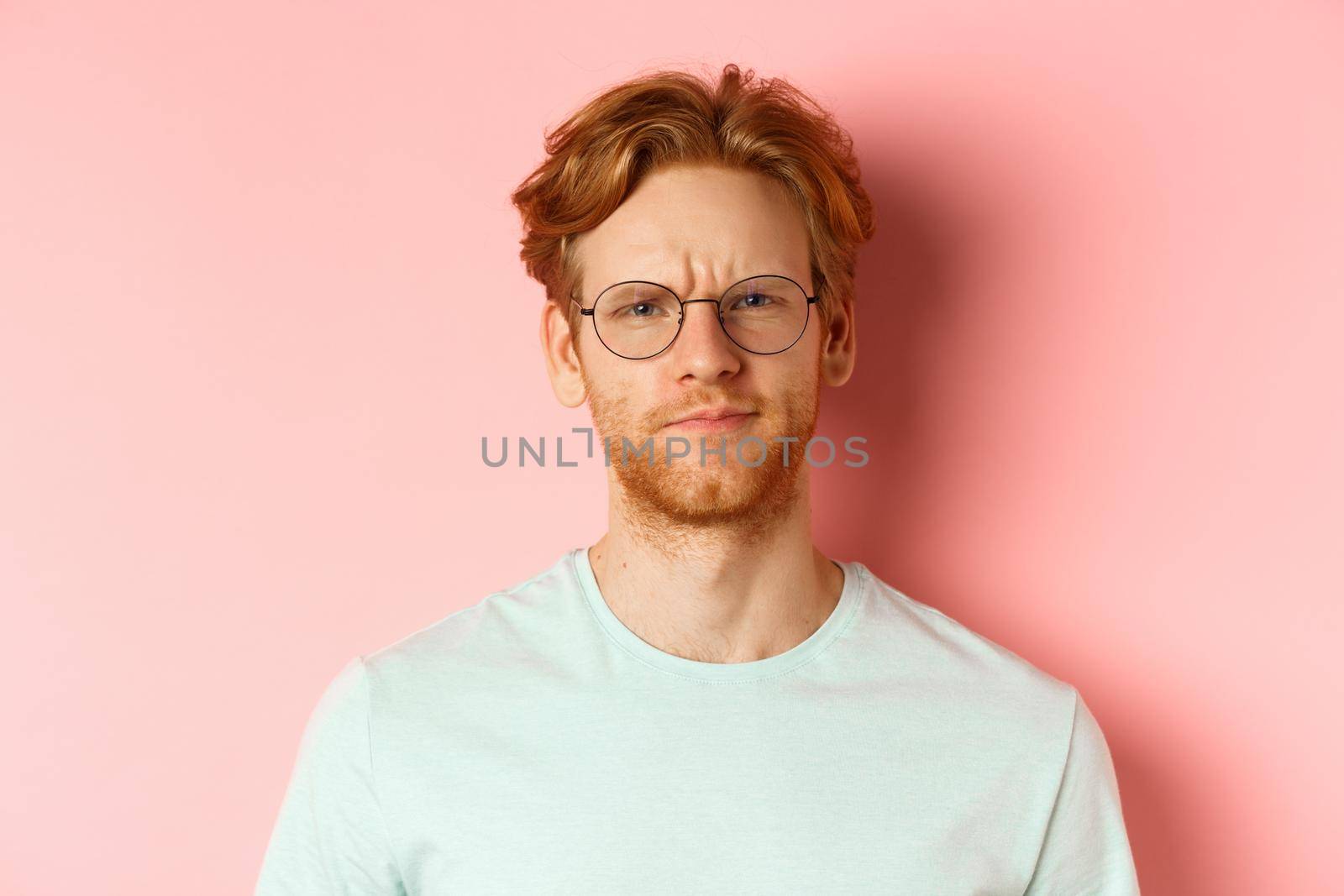Headshot of skeptical redhead man in glasses and t-shirt frowning disappointed, staring with disapproval and judgement, standing over pink background.