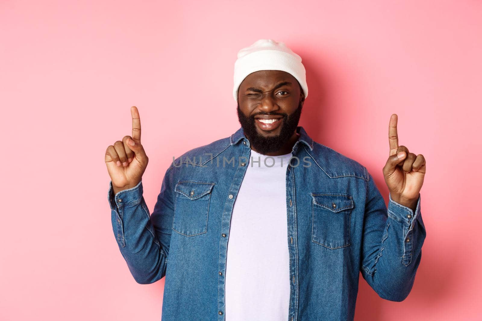 Cheeky african american man smiling and winking, showing deal on top, pointing fingers up, standing over pink background.