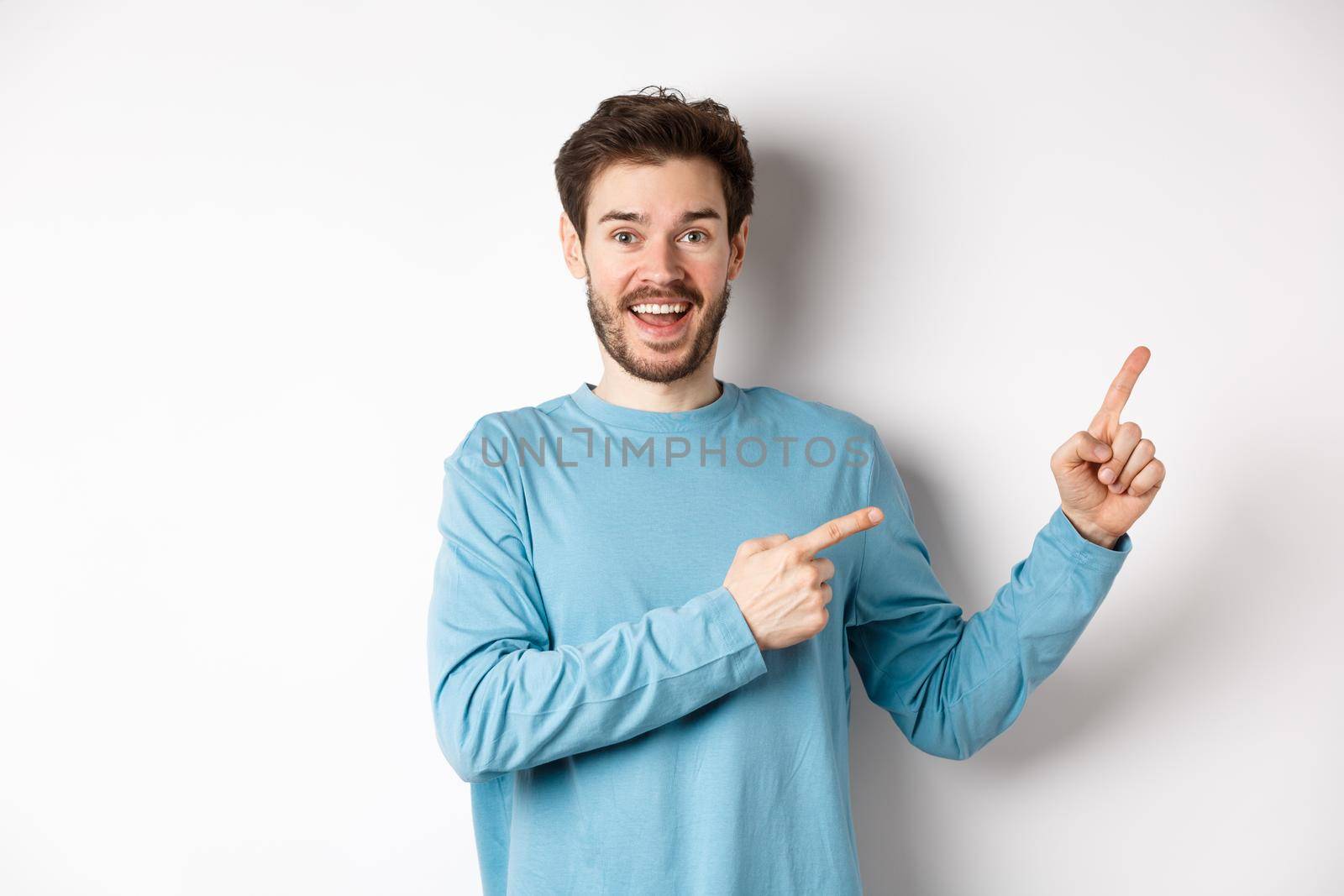 Cheerful smiling man pointing fingers right and showing banner or logo on white space for advertisements, recommending product, standing over studio background by Benzoix