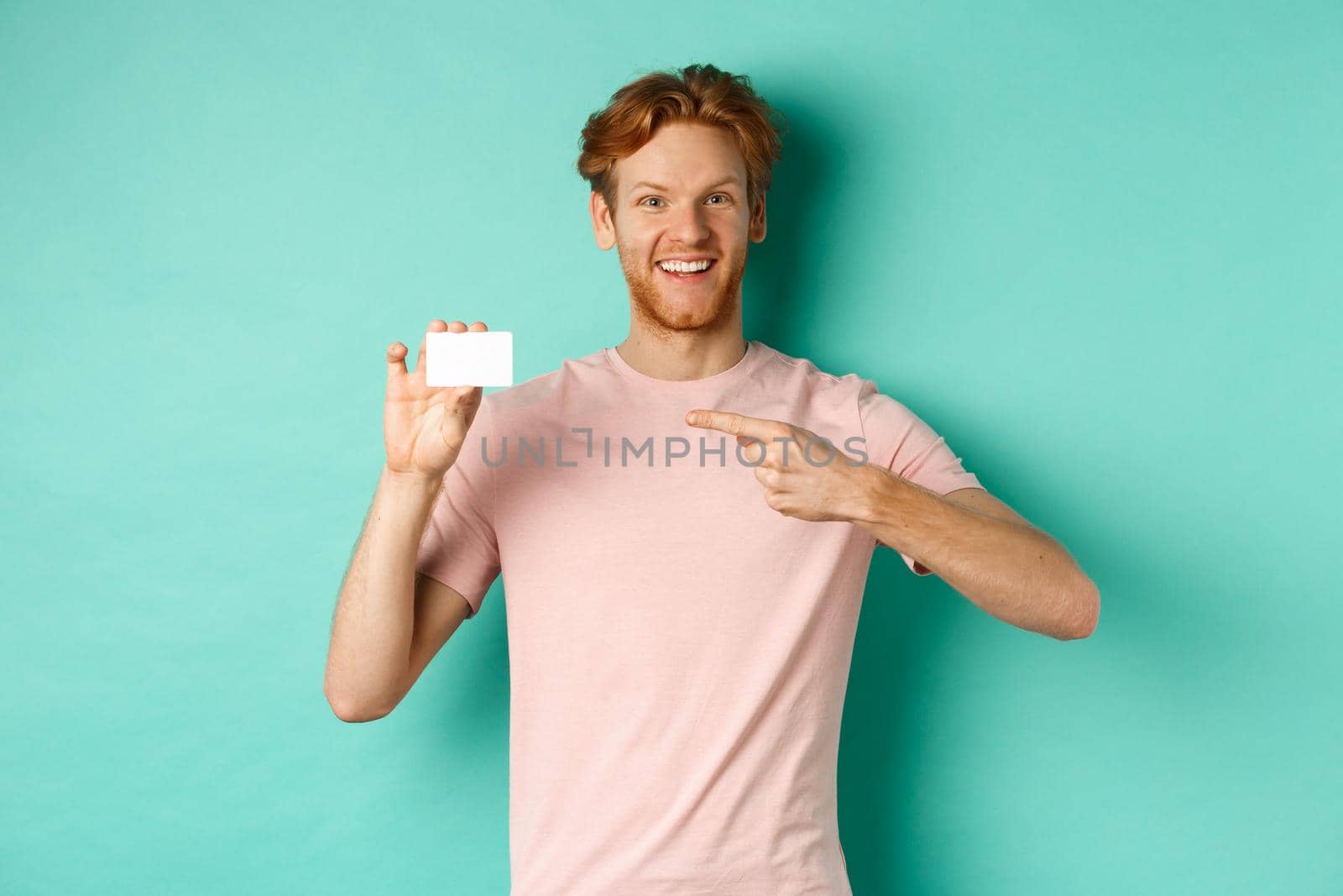 Attractive adult man with beard and red hair pointing finger at plastic credit card, smiling pleased at camera, standing over turquoise background by Benzoix