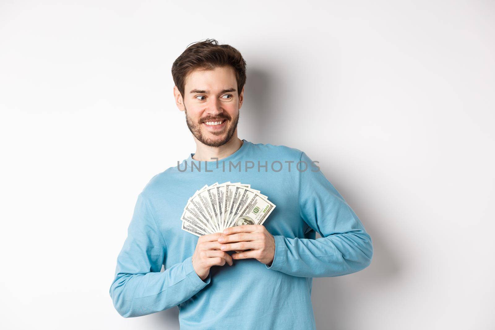 Greedy smiling man showing money and looking right, thinking about shopping, standing over white background by Benzoix