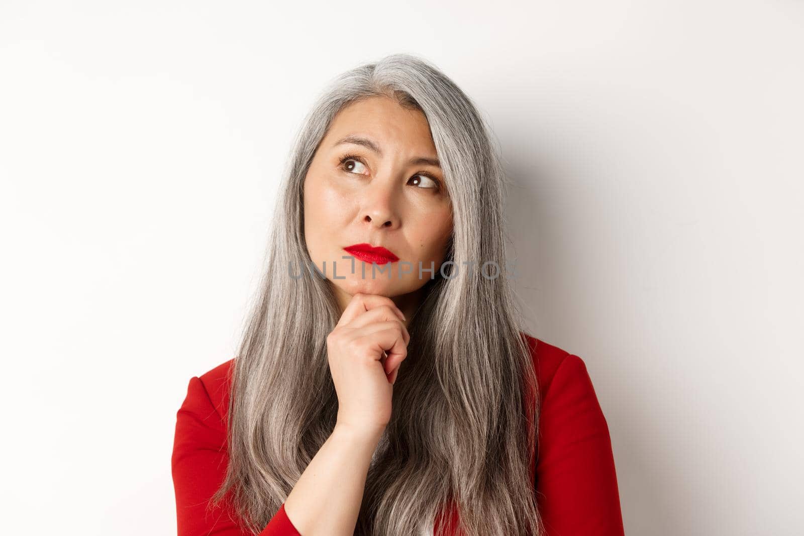 Close up of thoughtful asian business woman looking at upper left corner, thinking or making choice, standing over white background.