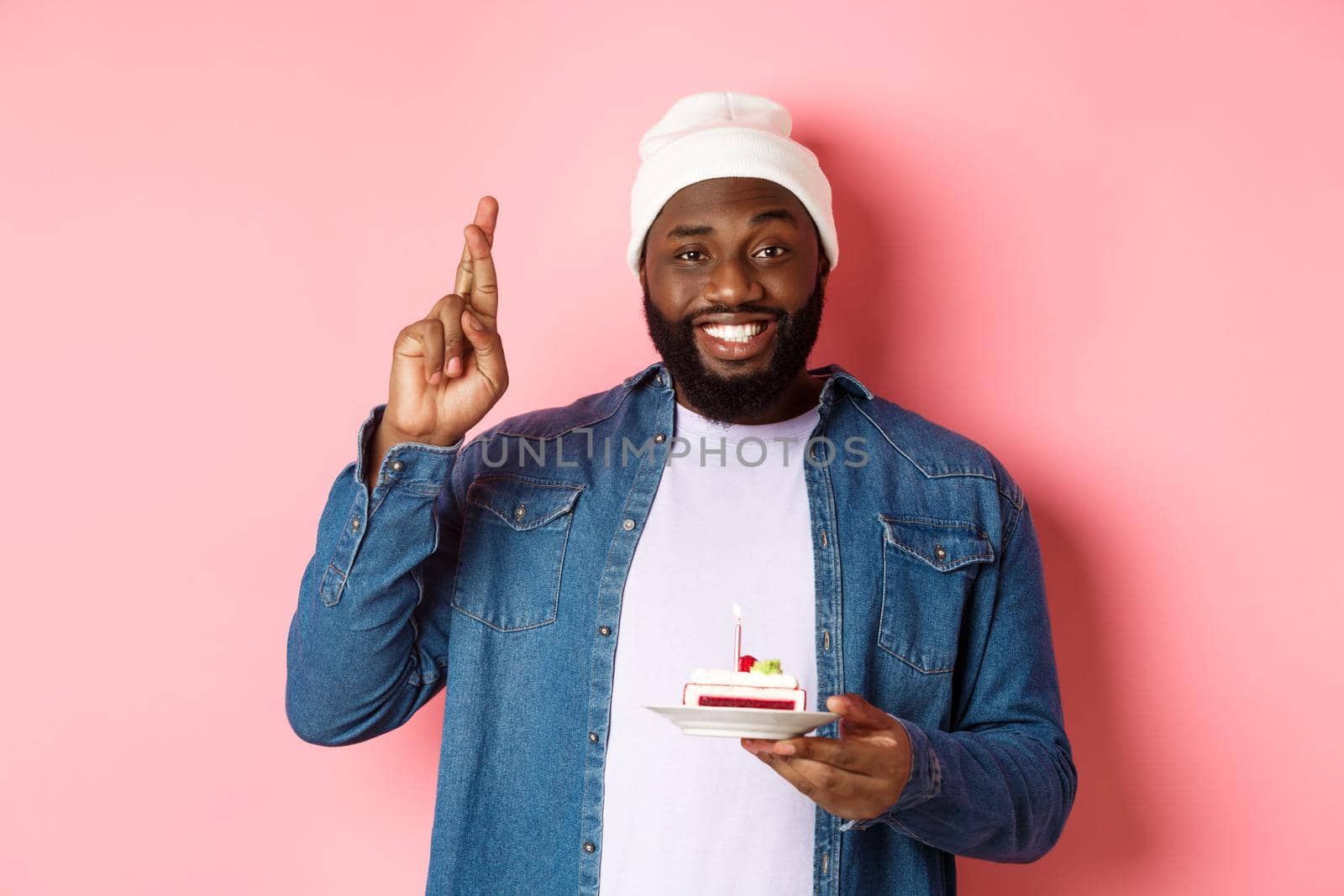 Handsome african-american guy celebrating birthday, making wish with fingers crossed, holding bday cake with candle, standing against pink background by Benzoix