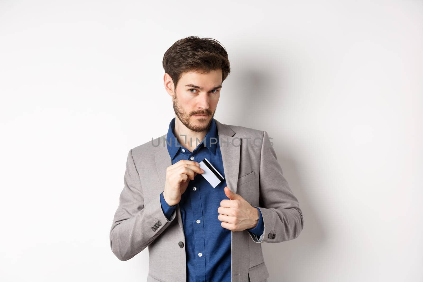 Confident and successful businessman looking self-assured while put plastic credit card in suit pocket, standing on white background by Benzoix