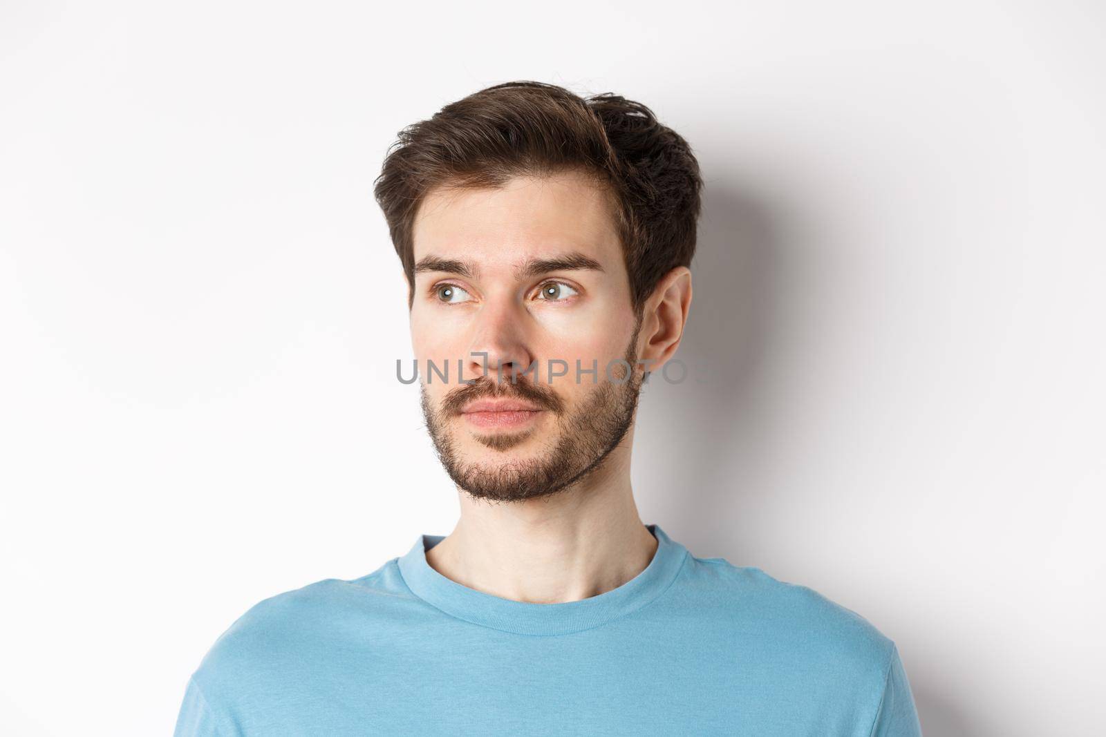 Close-up of handsome male model with beard and moustache, looking left with thoughtful face, standing over white background.