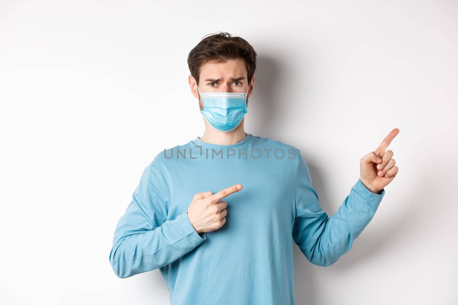 Coronavirus, health and quarantine concept. Sad man in medical mask frowning, pointing fingers right with doubtful face, standing over white background by Benzoix