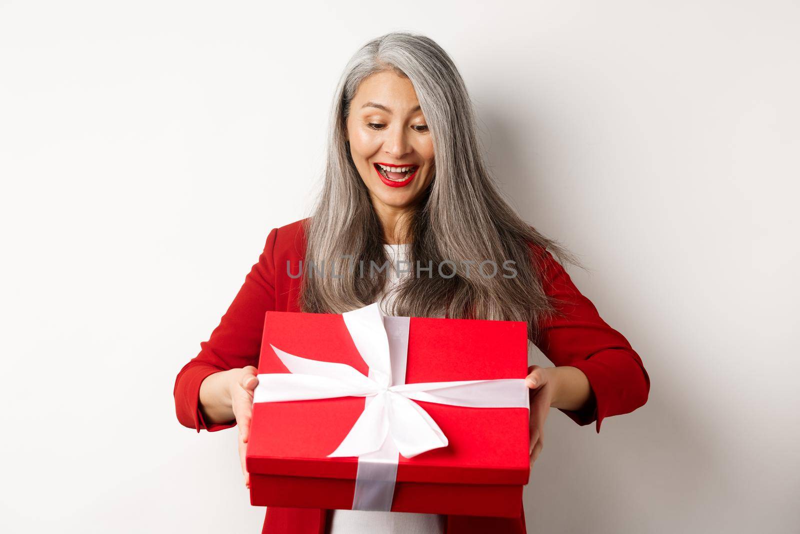 Happy elderly woman with grey hair, receive present, looking at red gift box and smiling surprised, standing over white background by Benzoix