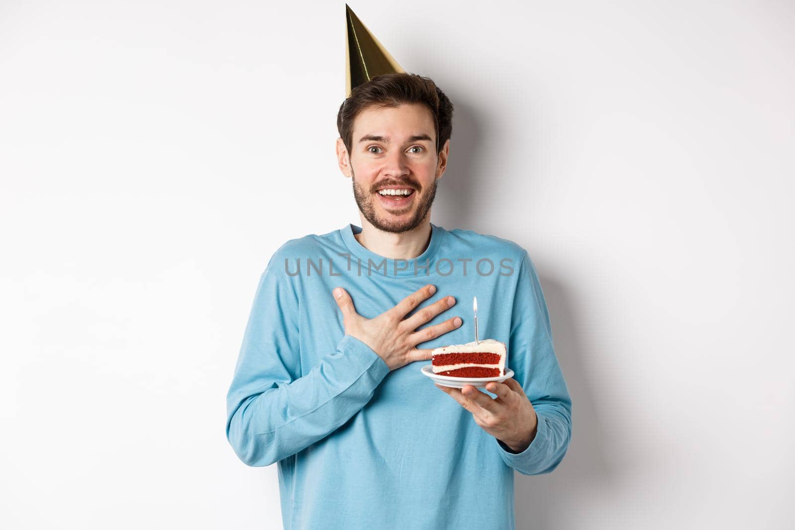 Celebration and holidays concept. Surprised birthday boy in party hat, holding bday cake and looking thankful, standing over white background by Benzoix