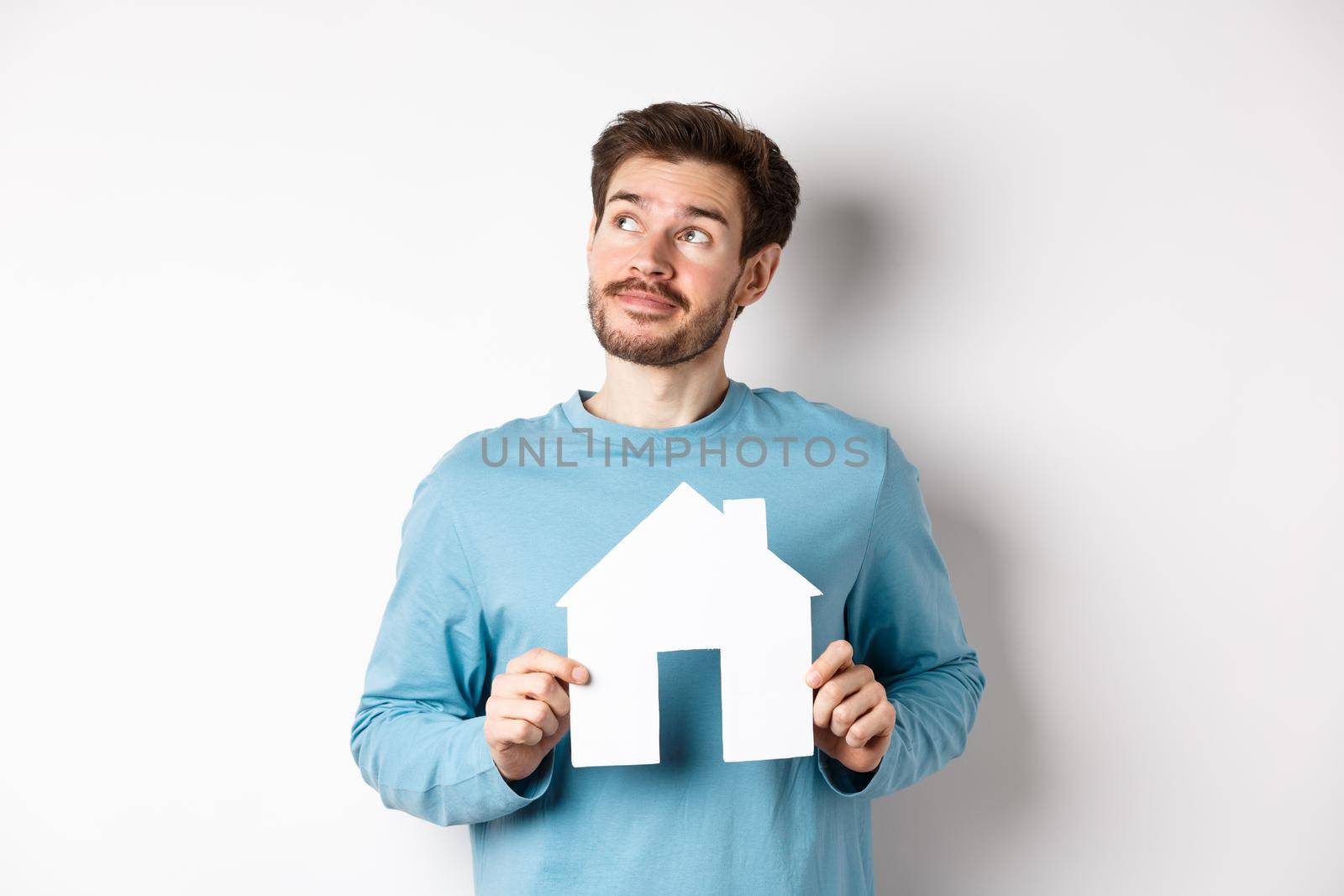 Real estate and insurance concept. Man dreaming about buying property, holding paper house cutout and looking at upper left corner, imaging things, white background by Benzoix