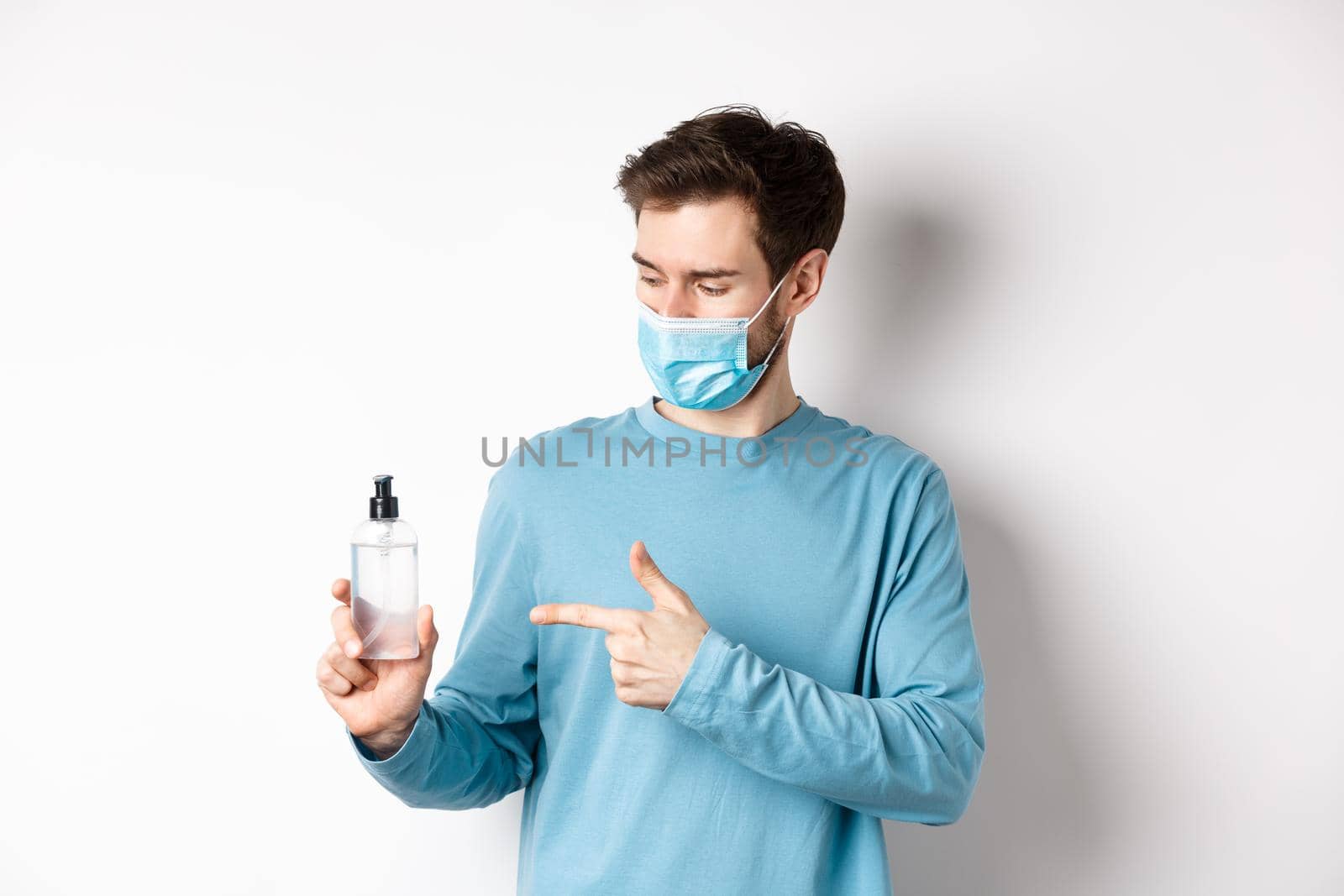 Covid-19, health and quarantine concept. Young man pointing and looking at hand sanitizer bottle, showing good antiseptic, white background by Benzoix