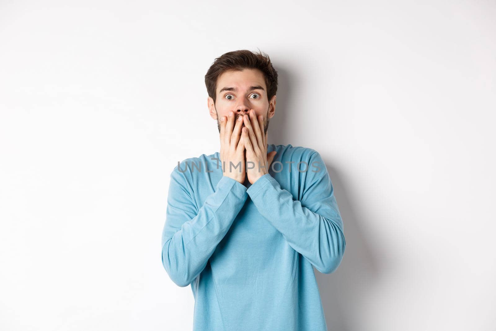 Shocked and startled cauacasian man, gasping and covering mouth with hands, staring at something scary, standing over white background by Benzoix