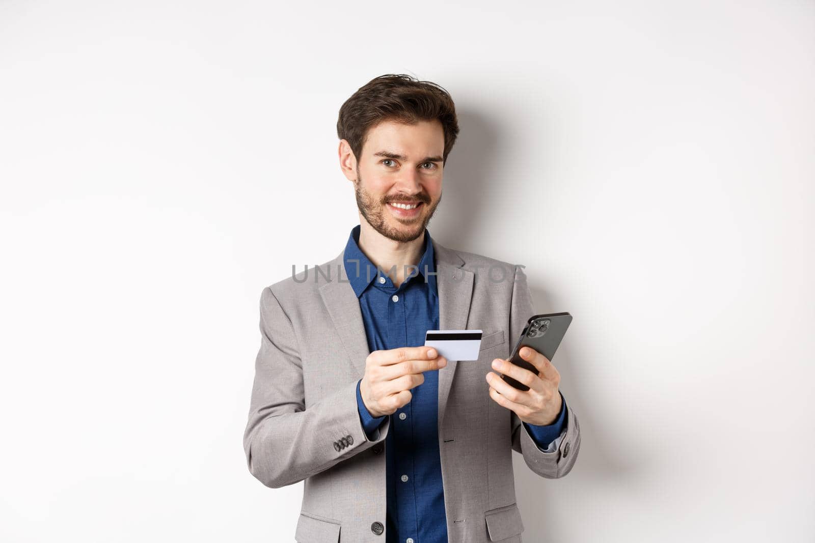 Online shopping. Handsome businessman in suit paying with credit card on smartphone, smiling satisfied at camera, standing on white background.