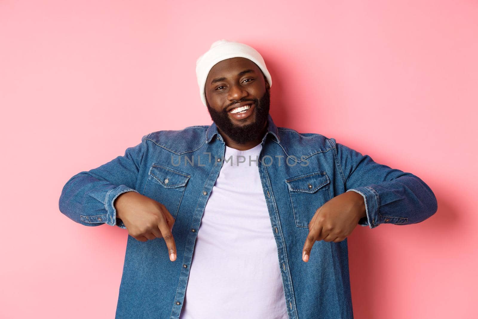 Happy Black man with beard, wearing hipster clothes, pointing fingers down and smiling, showing promo offer copy space, pink background.