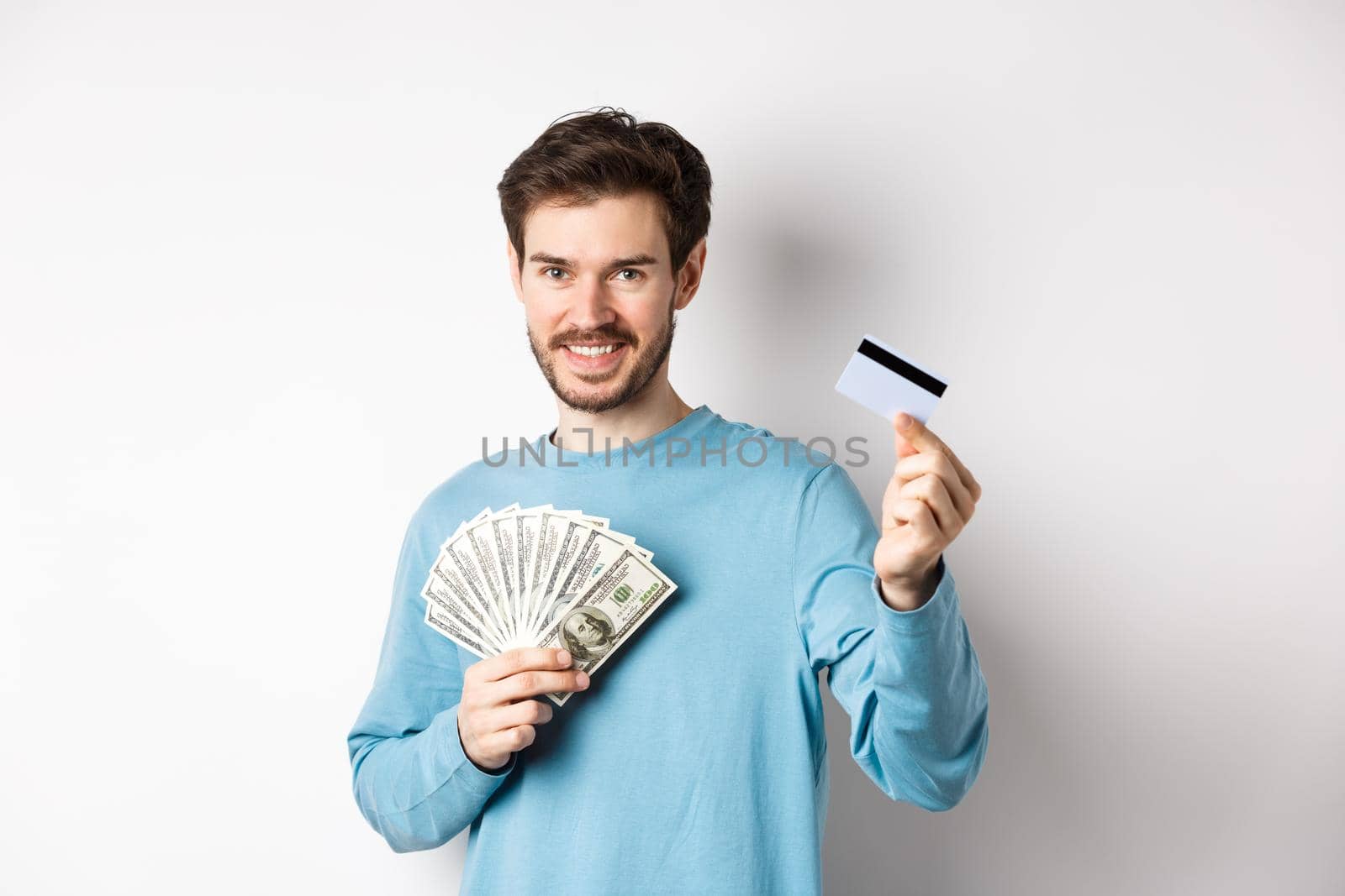 Handsome young man smiling and offering payment in cash and contactless, showing money with plastic credit card, standing on white background by Benzoix