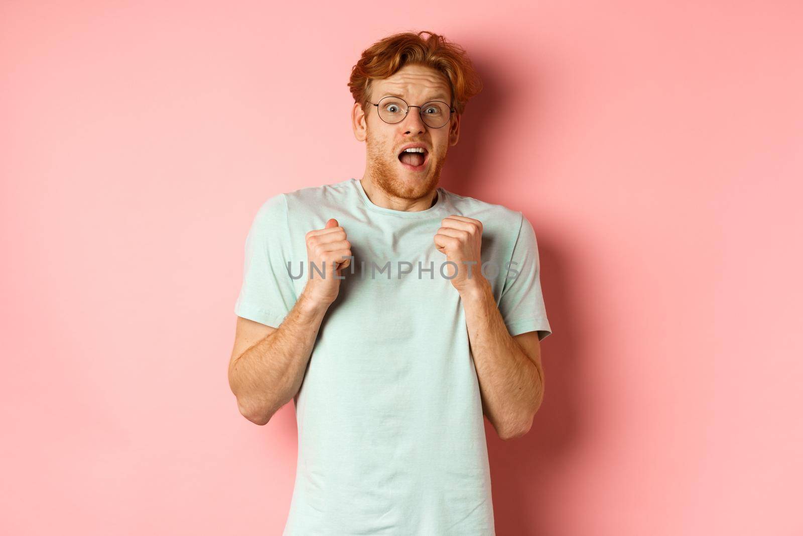 Portrait of scared redhead guy in glasses staring startled at camera, press hands to body and scream of fear, standing over pink background by Benzoix