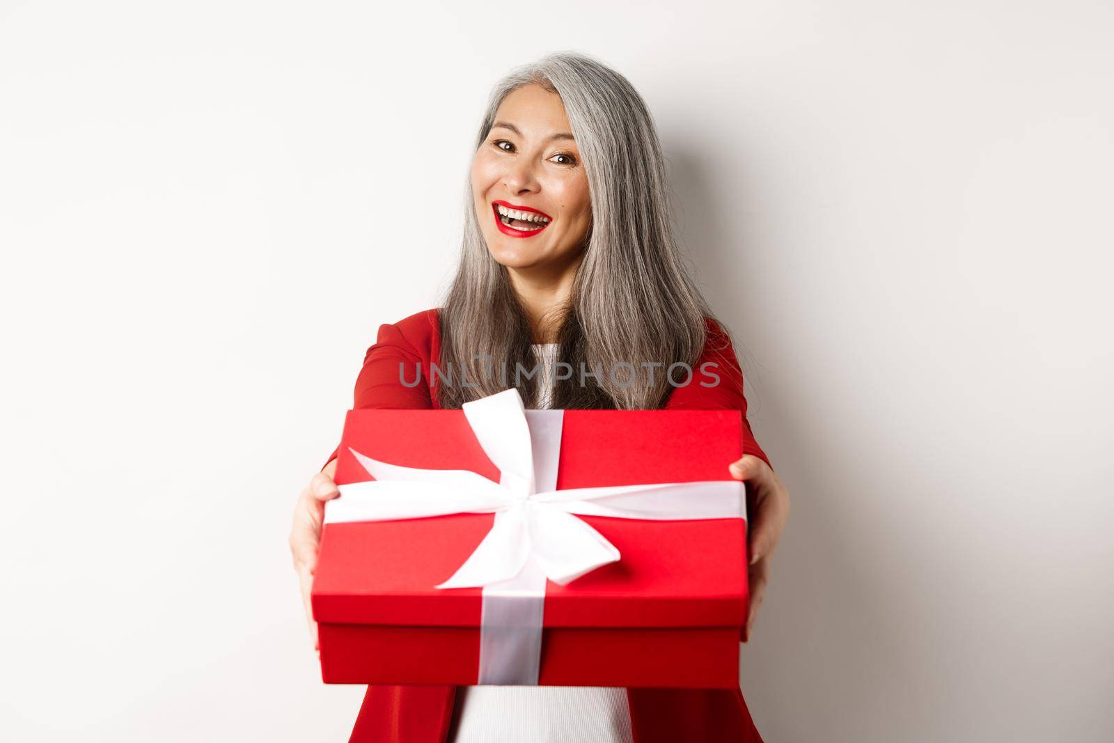 Happy holidays concept. Cheerful asian senior woman giving you red gift box and smiling, congratulating with valentine day, standing over white background by Benzoix