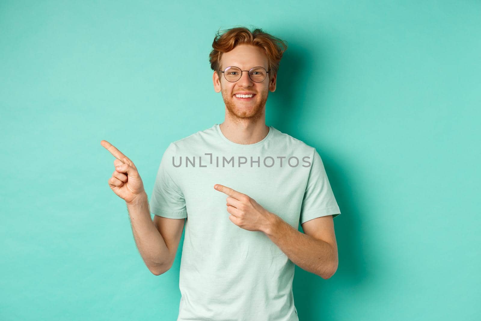 Young cheerful man with short red hair and glasses, pointing fingers left at copy space, smiling white teeth, showing advertisement, mint background by Benzoix