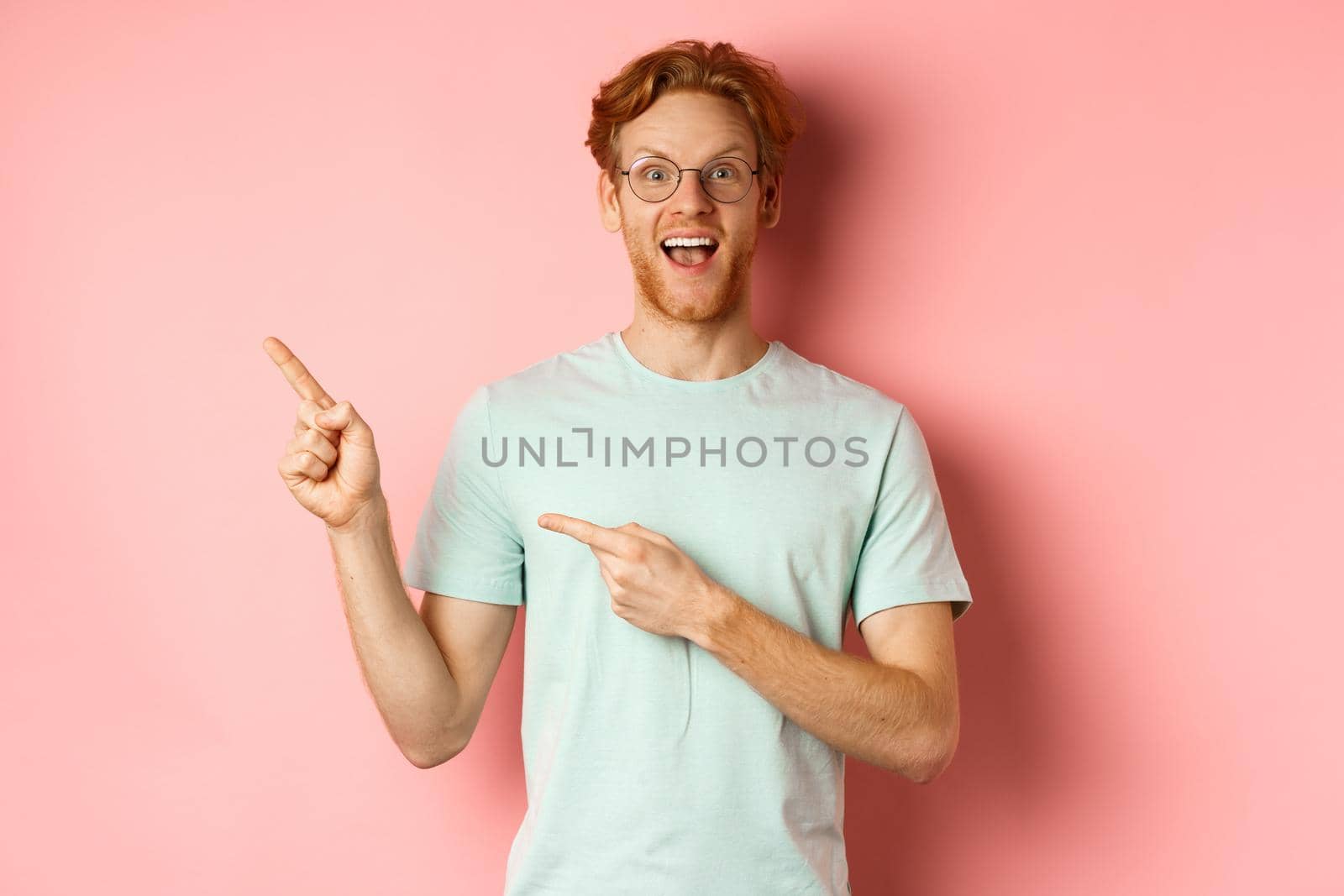 Portrait of cheerful young man with red hair, wearing glasses, pointing fingers at upper left corner and smiling, standing over pink background.