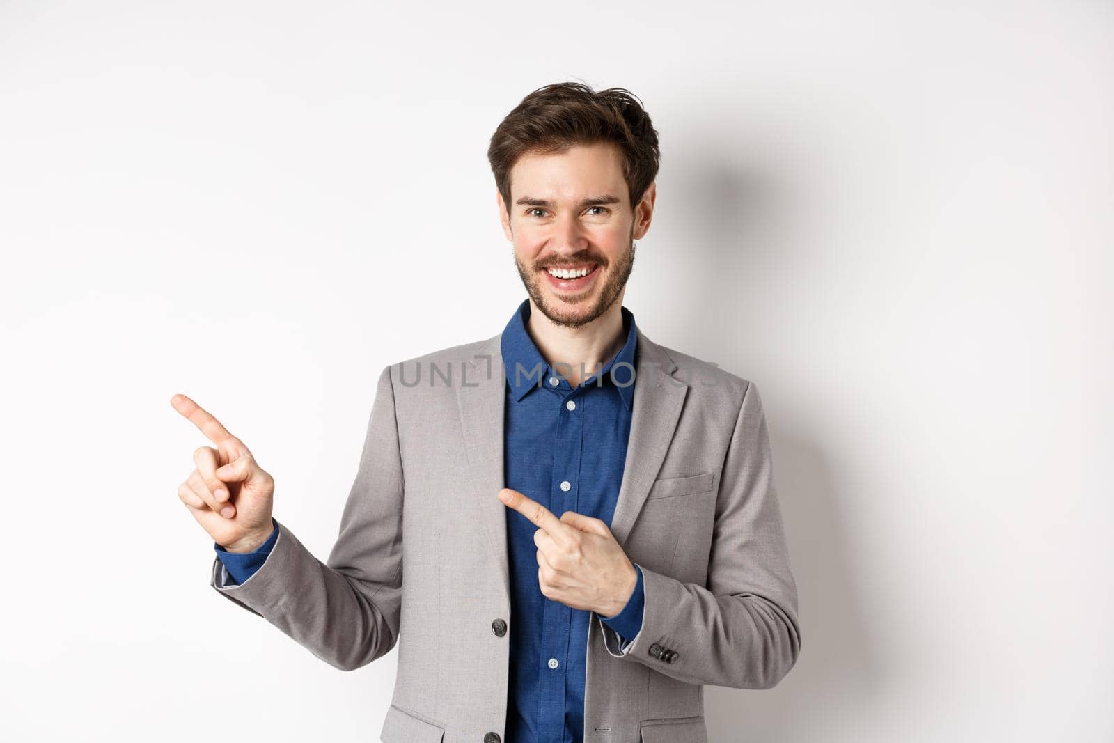 Handsome caucasian man in business suit pointing fingers left at banner and smiling, showing advertisement, standing against white background by Benzoix