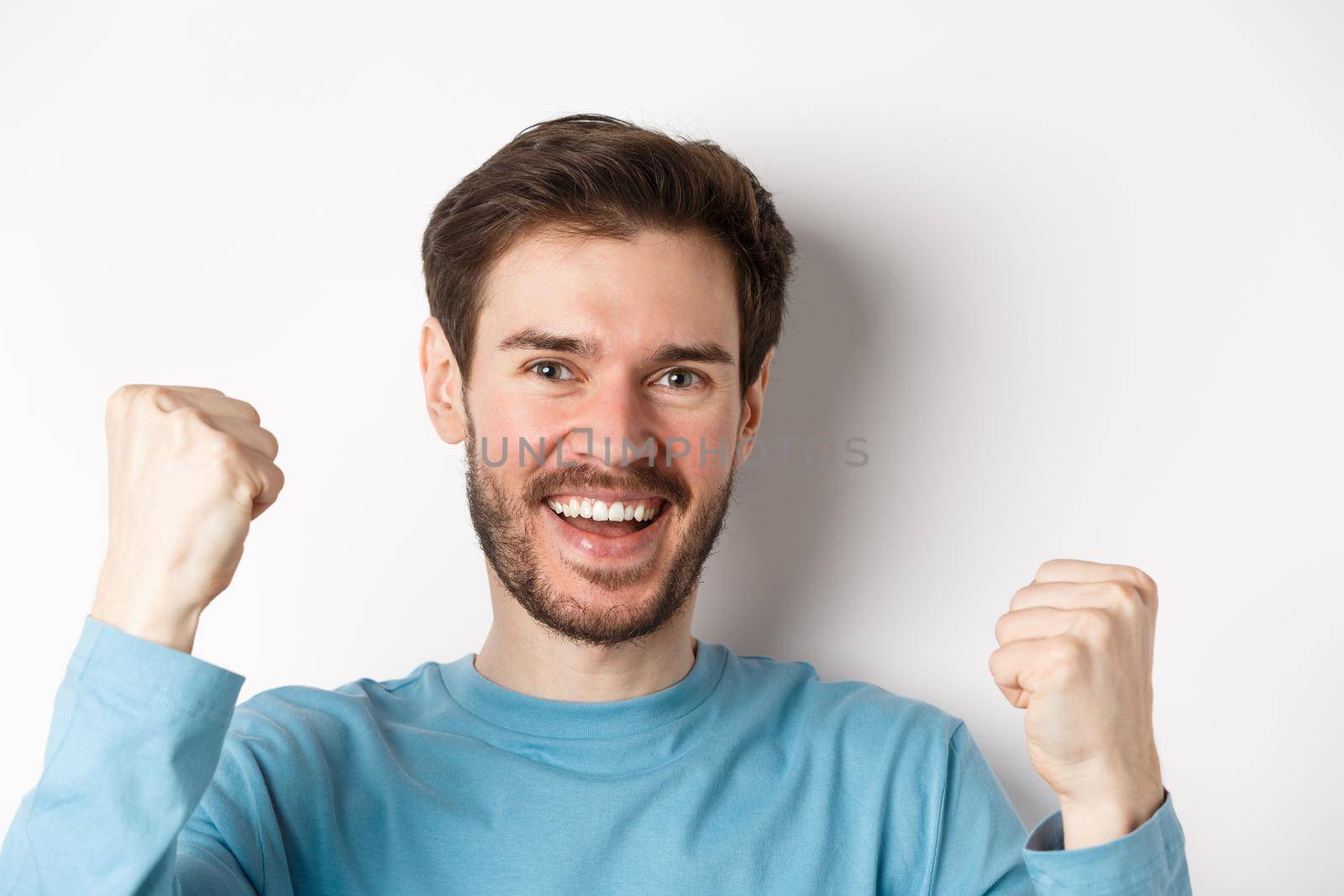 Close-up of happy young man celebrating, winning prize and say yes, smiling satisfied with fist pumps, standing over white background.