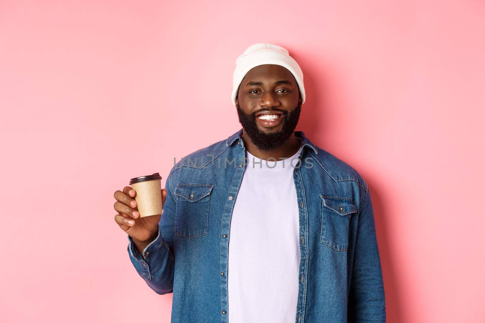 Handsome modern Black man drinking takeaway coffee, smiling and looking satisfied at camera, standing over pink background.