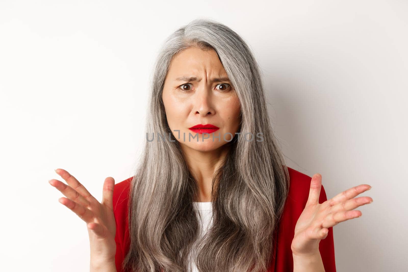 Close up of confused asian female manager with grey hair, wearing red blazer and makeup, spread hands sideways and staring puzzled at camera, white background by Benzoix
